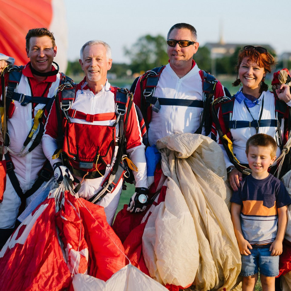 Group of skydivers in red and white jumpsuits smiling with parachutes at the Plano Balloon Festival.